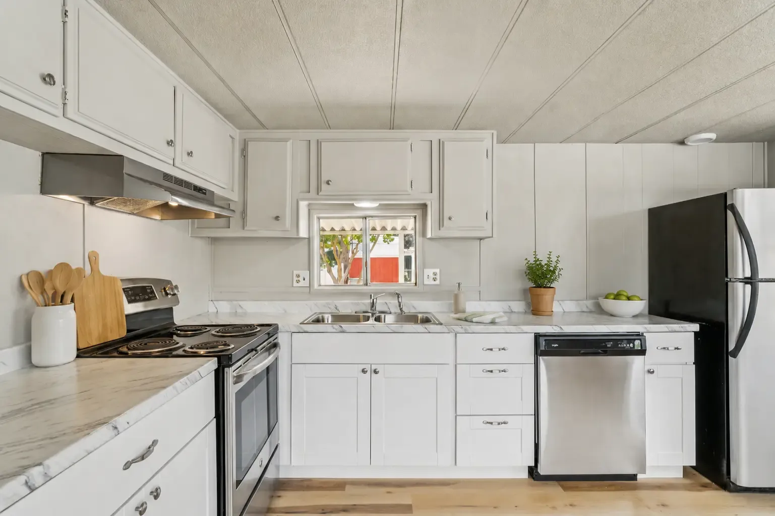 Muro de cocina con estufa y campana de acero inoxidable, fregadero doble bajo una ventana, cubiertas tipo mármol y gabinetes blancos
