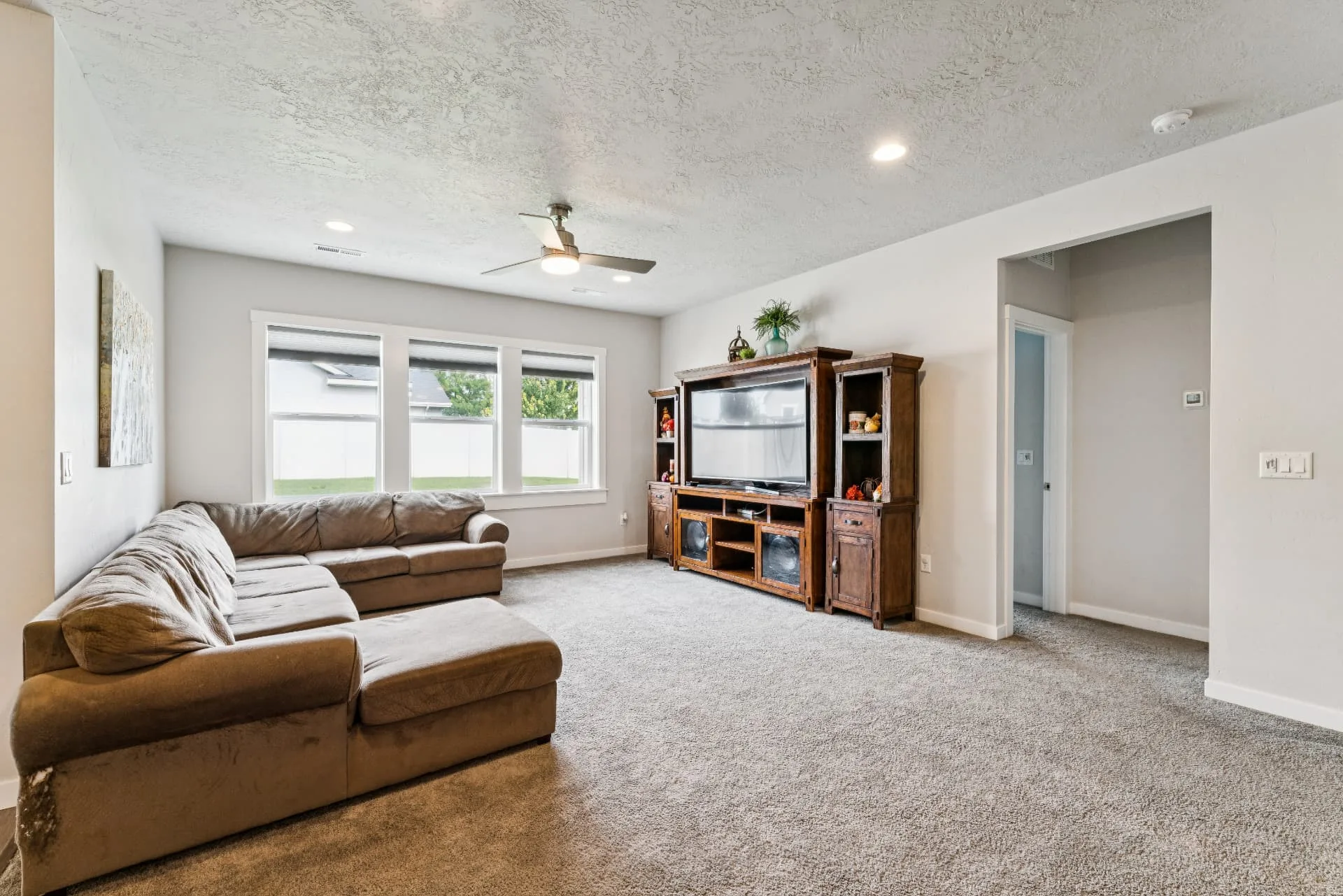 Carpeted living room with a ceiling fan, three windows facing the backyard and an open doorway to the hall