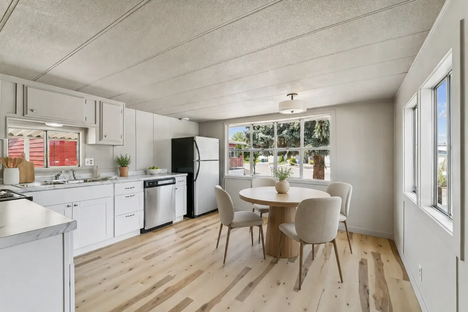 Cocina con gabinetes blancos, lavavajillas y refrigerador junto a un comedor con una ventana grande