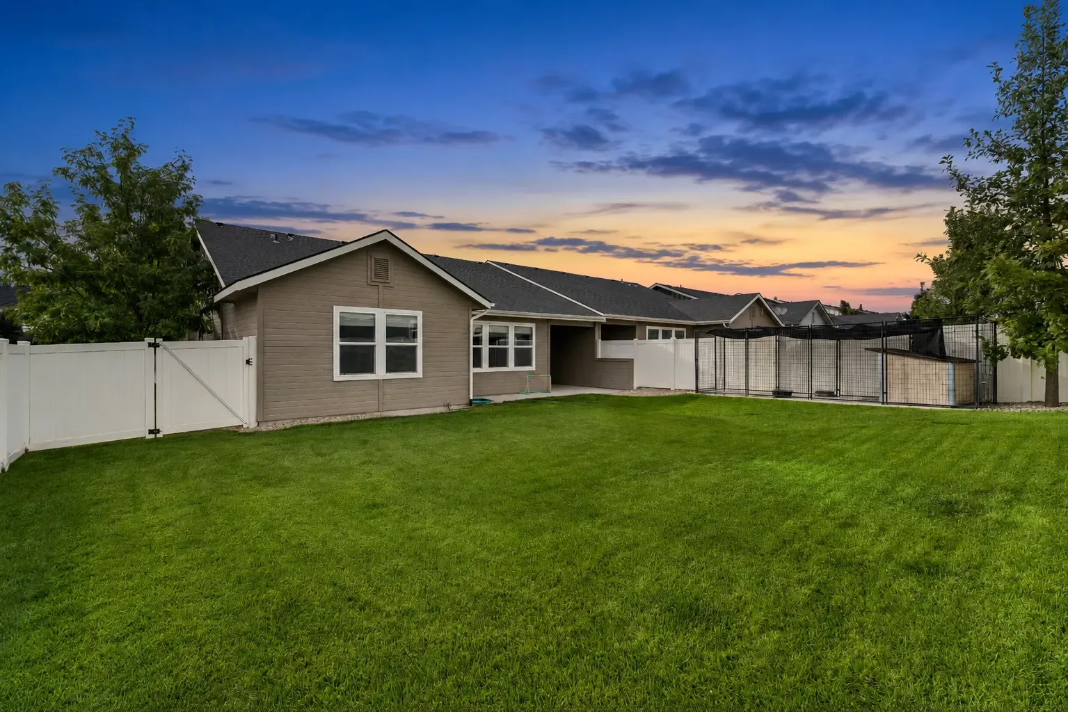 Fenced backyard at dusk with grass, mature trees and a fenced kennel area along the rear fence